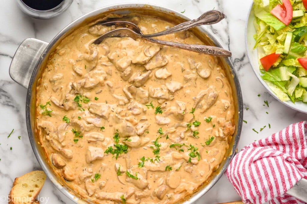 overhead of stainless steel skillet of Beef Stroganoff sauce with two spoons for serving. Bowl of lettuce salad, glass of wine, and piece of bread surrounding