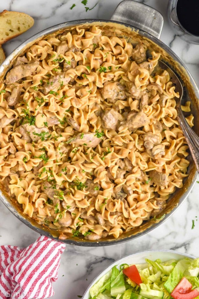 overhead of stainless steel skillet of Beef Stroganoff with two spoons for serving. Piece of bread, glass of red wine, and bowl of lettuce salad surrounding skillet