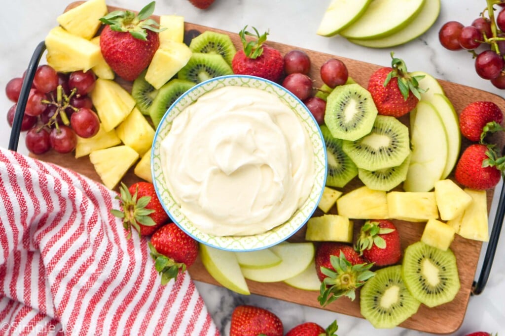 overhead of bowl of fruit dip on a serving board with fresh pineapple, strawberries, kiwi, and purple grapes