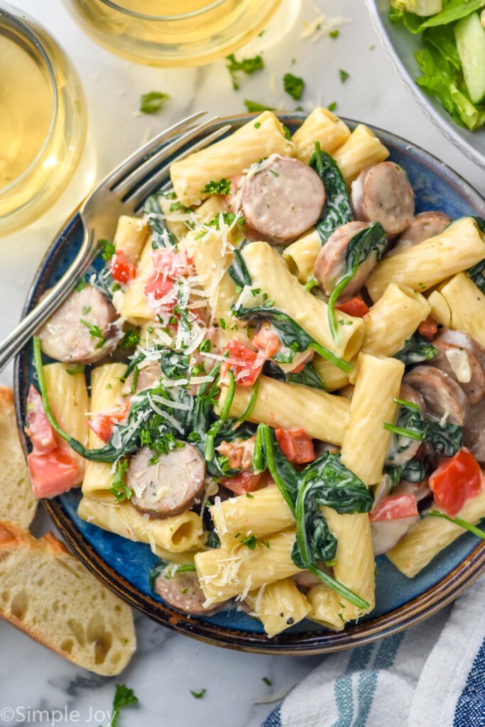 overhead of plate of One Pot Sausage Pasta topped with fresh parmesan cheese and a fork sitting beside. Two glasses of white wine, bread, and bowl of salad surrounding.