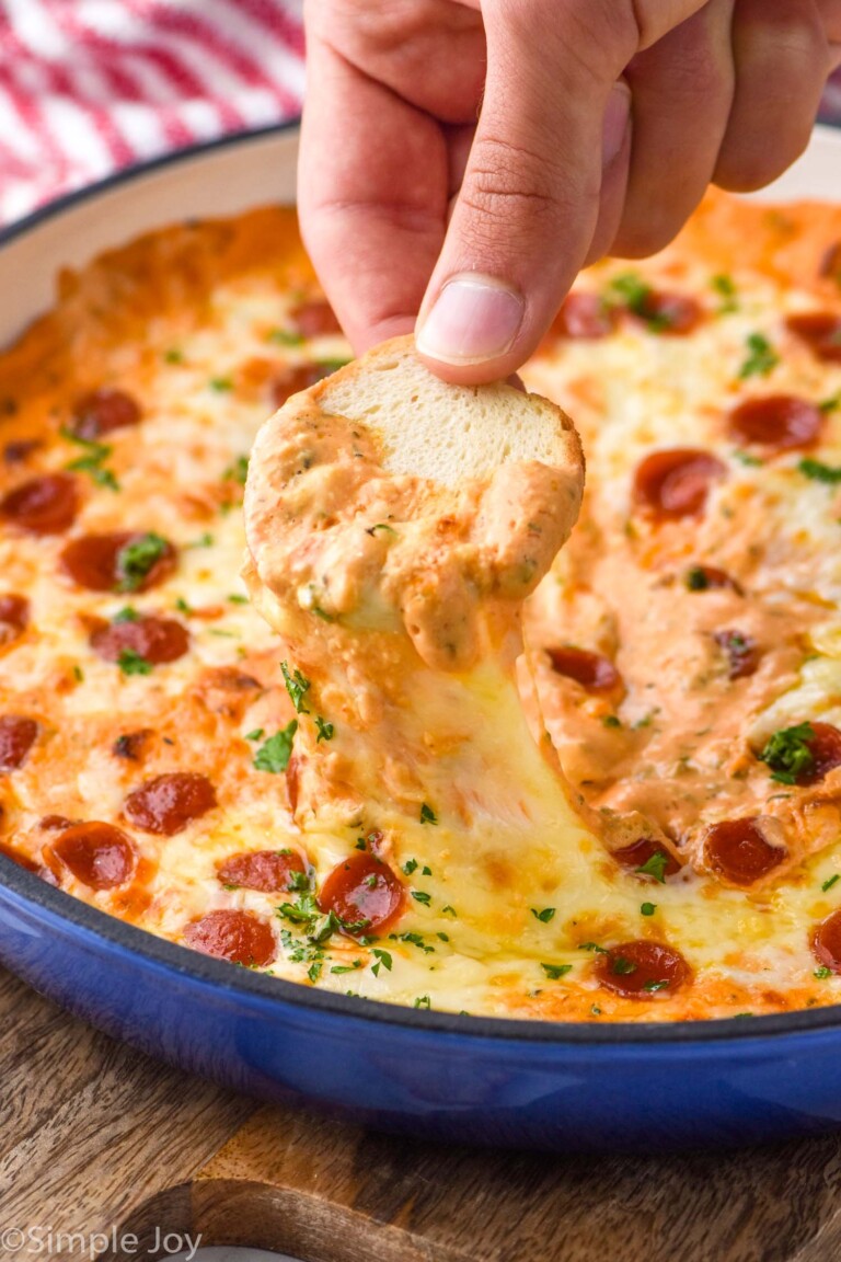 man's hand holding toasted baguette dipped in skillet of Pizza Dip topped with pepperoni and fresh parsley