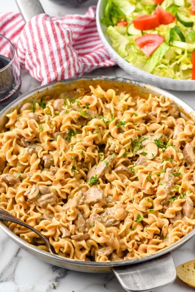 stainless steel skillet of Beef Stroganoff. Bowl of lettuce salad and glass of red wine sitting in background