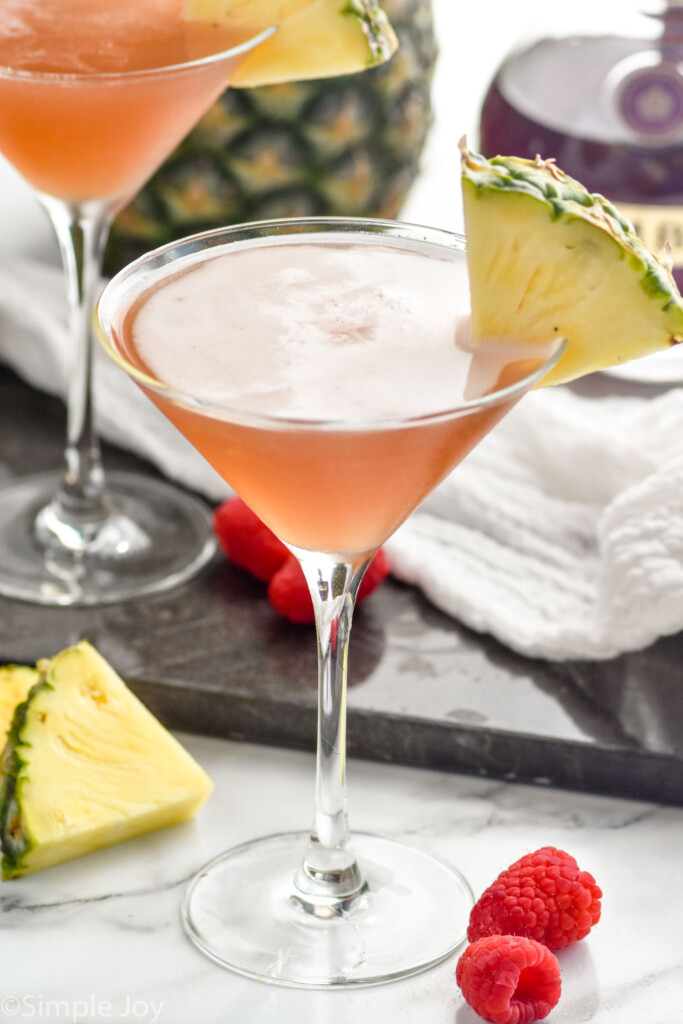 martini glass of french martini with a pineapple wedge on the rim. Fresh raspberries, pineapple slices, and french martini sitting in background