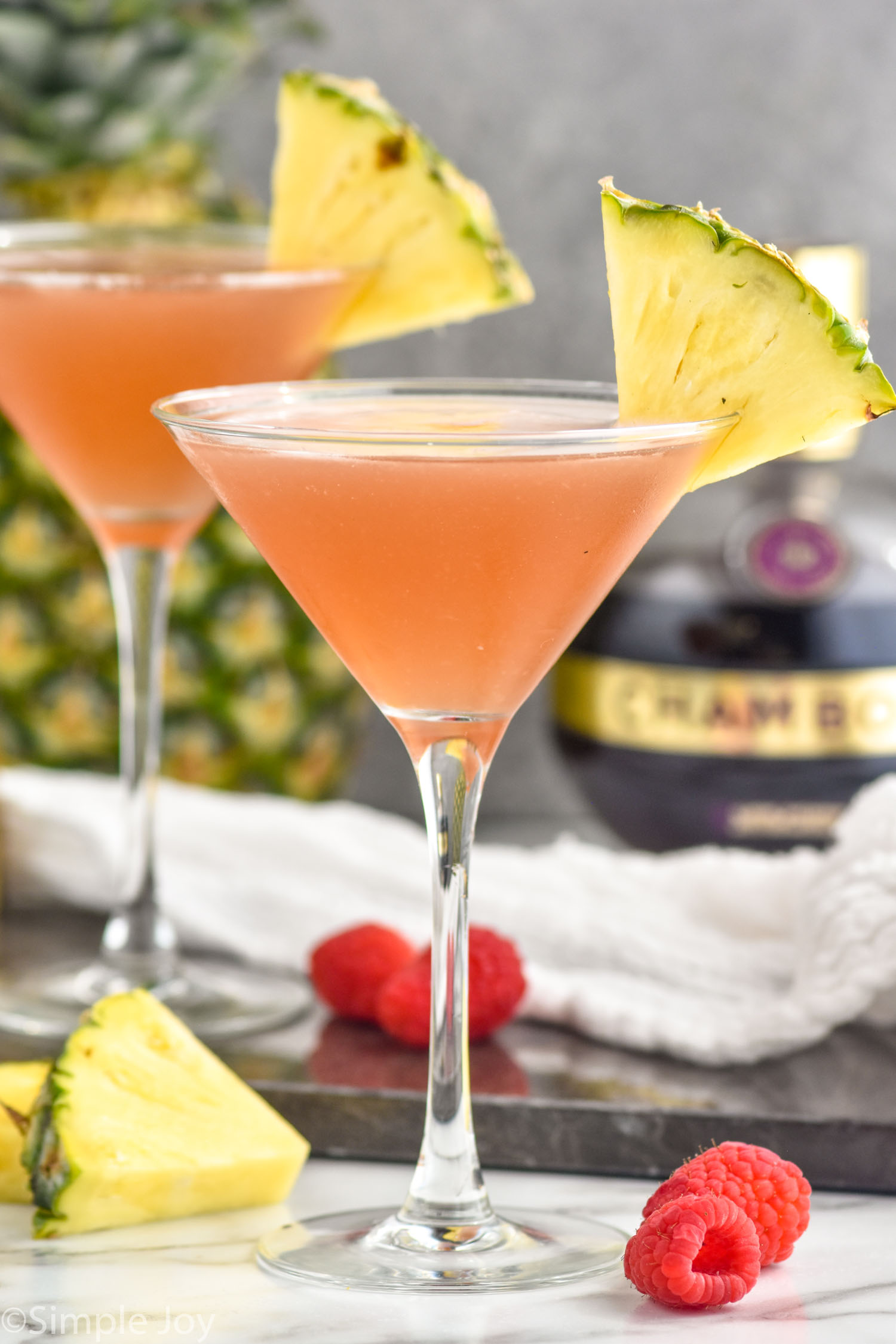 two glasses of French Martini garnished with a pineapple wedge. Fresh pineapple, raspberries, and bottle of Chambord liqueur sitting in background