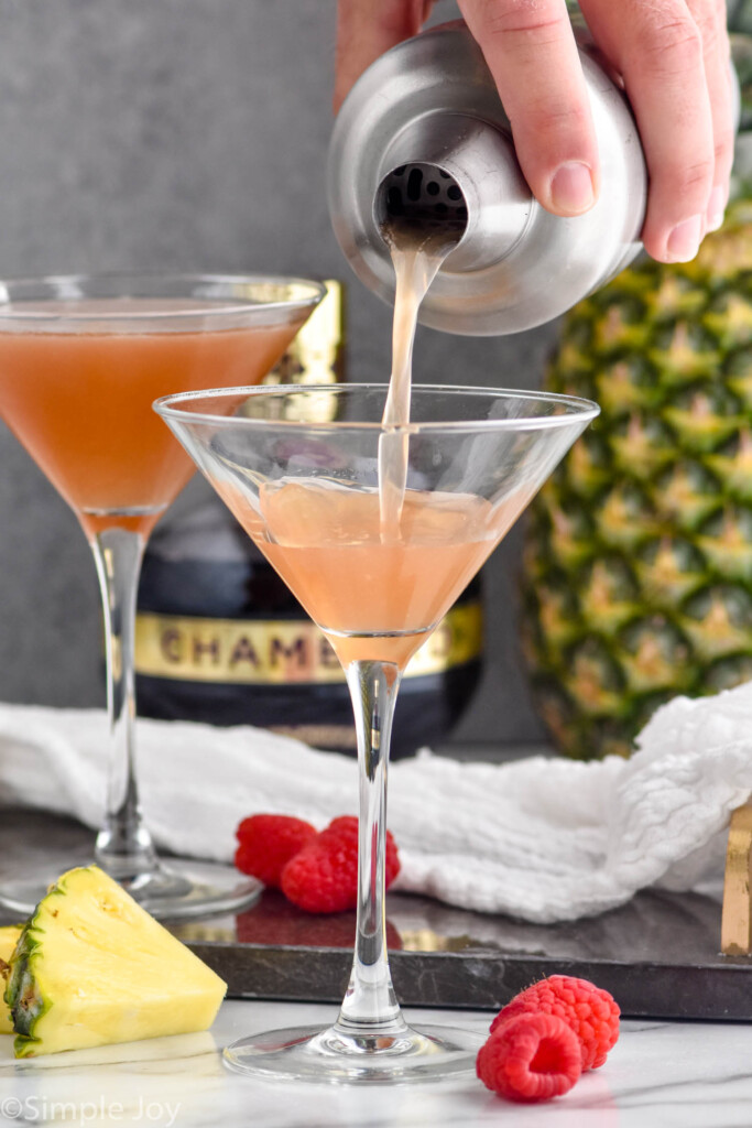 man's hand pouring cocktail shaker of French Martini ingredients pouring into a martini glass. Fresh pineapple wedge, raspberries, pineapple, and bottle of Chambord sitting beside and behind