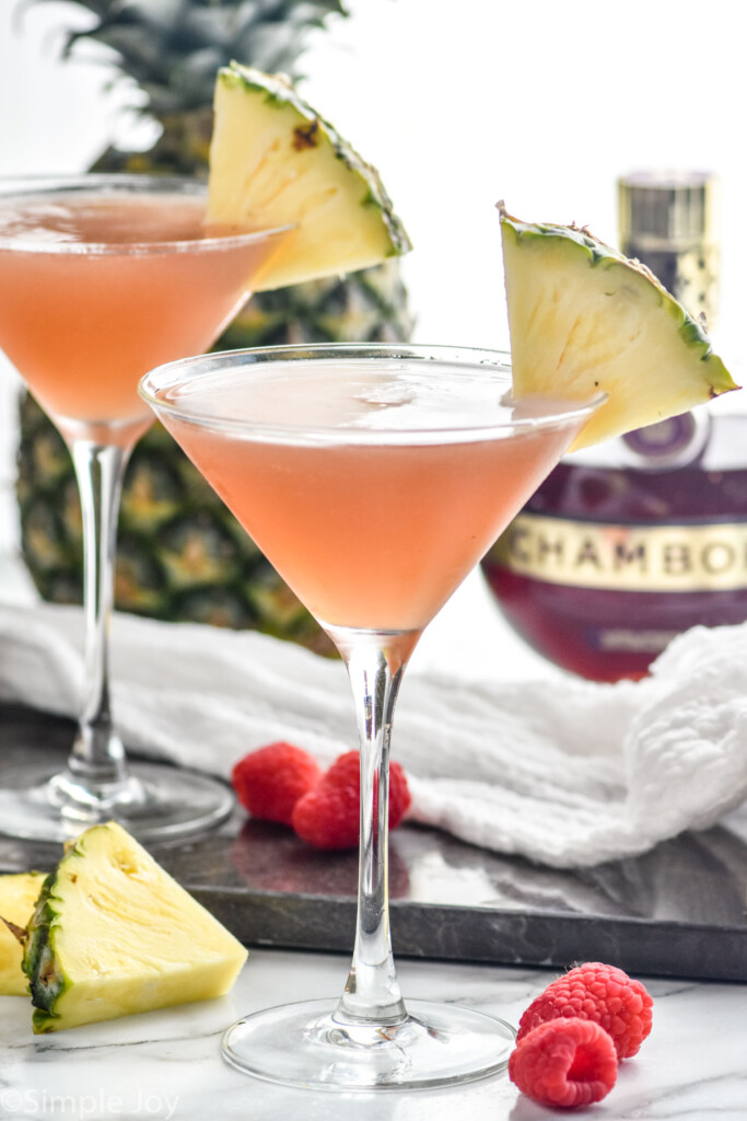 Two martini glasses of french martini garnished with a pineapple wedge. Fresh pineapple wedges, raspberries, and bottle of chambord sitting in background.