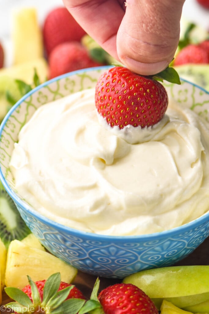 man's hand dipping fresh strawberry into a bowl of Fruit Dip. Fresh pineapple, apple slices, and strawberries sitting beside