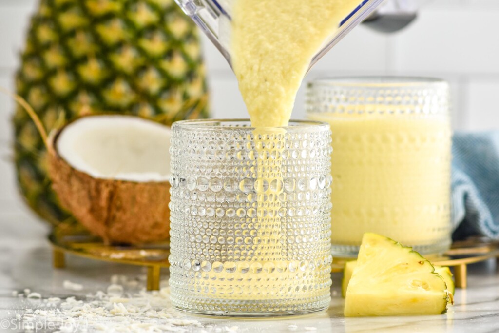 blender pouring Pina Colada Smoothie into a glass. Pineapple wedges and shredded coconut sitting beside. Glass of Pina Colada Smoothie, whole pineapple, and fresh coconut sitting in background