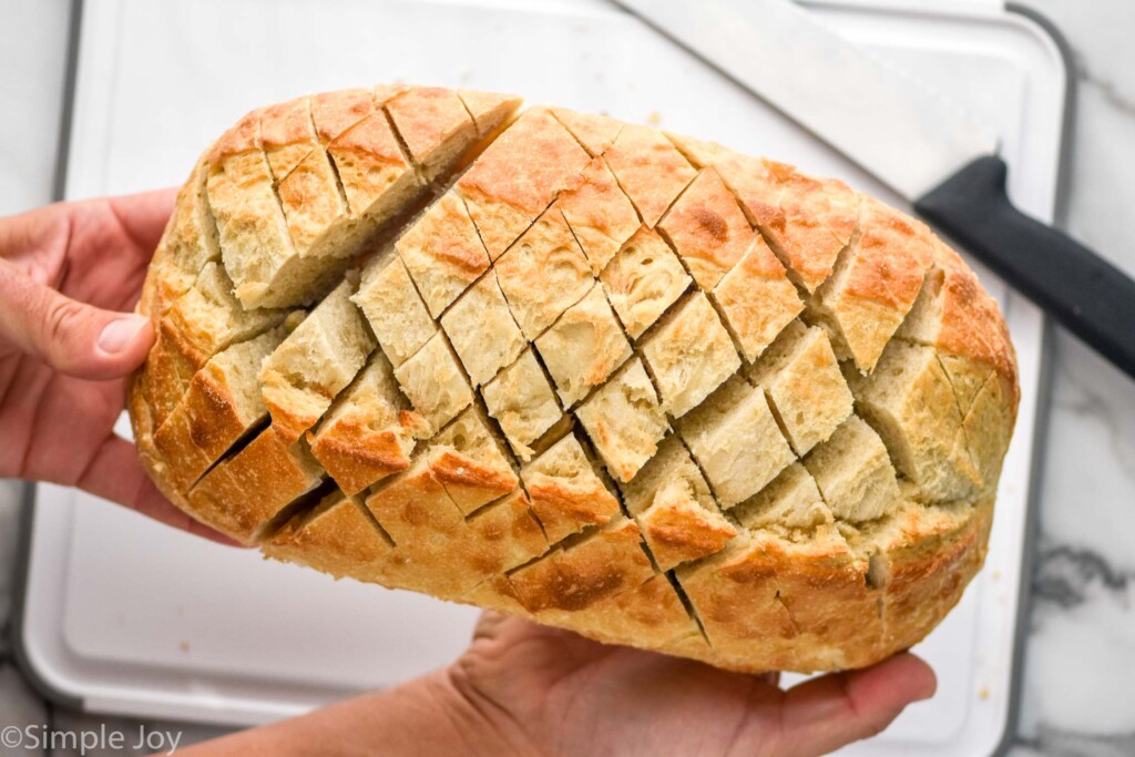 man's hands holding loaf of bread that has been cut to make Pull Apart Bread. Cutting board and knife underneath