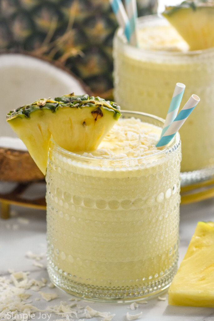 Glass of Pina Colada Smoothie topped with shredded coconut, garnished with a pineapple wedge and two straws. Shredded coconut and pineapple sitting beside