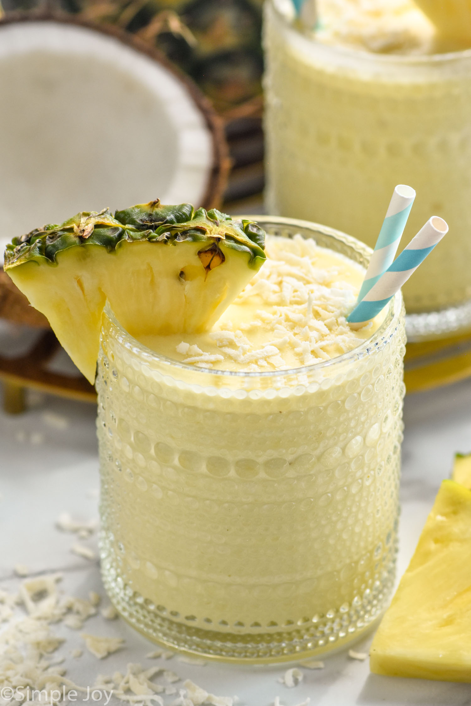 Glass of Pina Colada Smoothie topped with shredded coconut, garnished with a pineapple wedge and two straws.