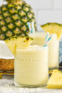 Two glasses of Pina Colada Smoothie topped with shredded coconut, pineapple wedge on the rim and two straws in the glass. Fresh pineapple sitting in background