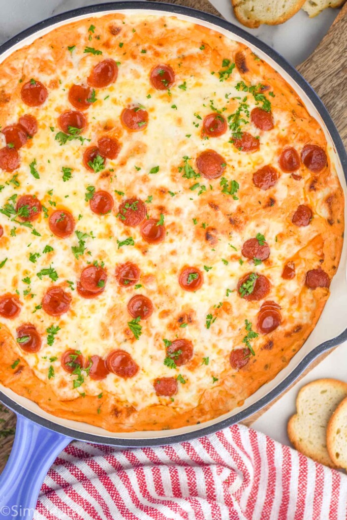 overhead of skillet of pizza dip topped with pepperoni and fresh parsley. Pieces of toasted bread sitting beside for serving