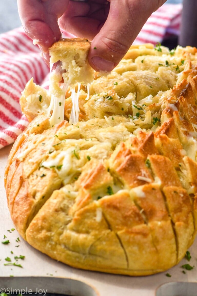 Man's hand pulling piece of Pull Apart Bread from loaf that is topped with melted cheese and fresh parsley