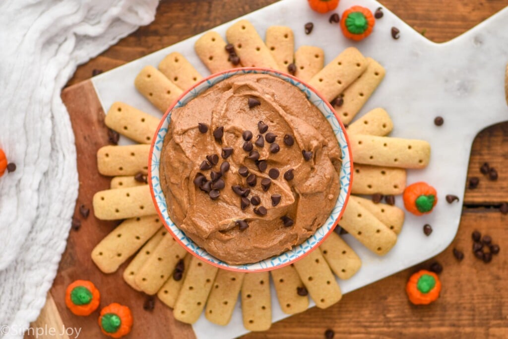 Overhead view of a bowl of Pumpkin Nutella Dip garnished with mini chocolate chips. Dipping sticks, candy pumpkins, and mini chocolate chips beside bowl on serving board.