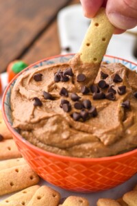 Person's hand dipping cracker into bowl of Pumpkin Nutella Dip garnished with mini chocolate chips. More dipping crackers beside bowl.