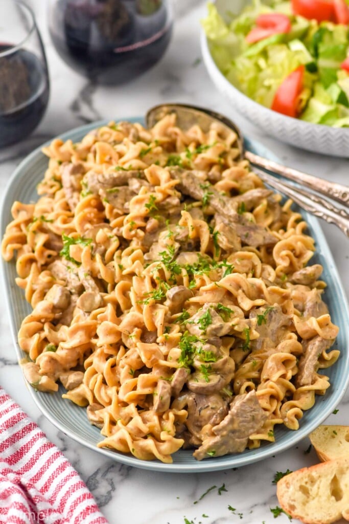 plate of Beef Stroganoff with spoons for serving. Pieces of bread, bowl of salad, and two glasses of red wine surrounding