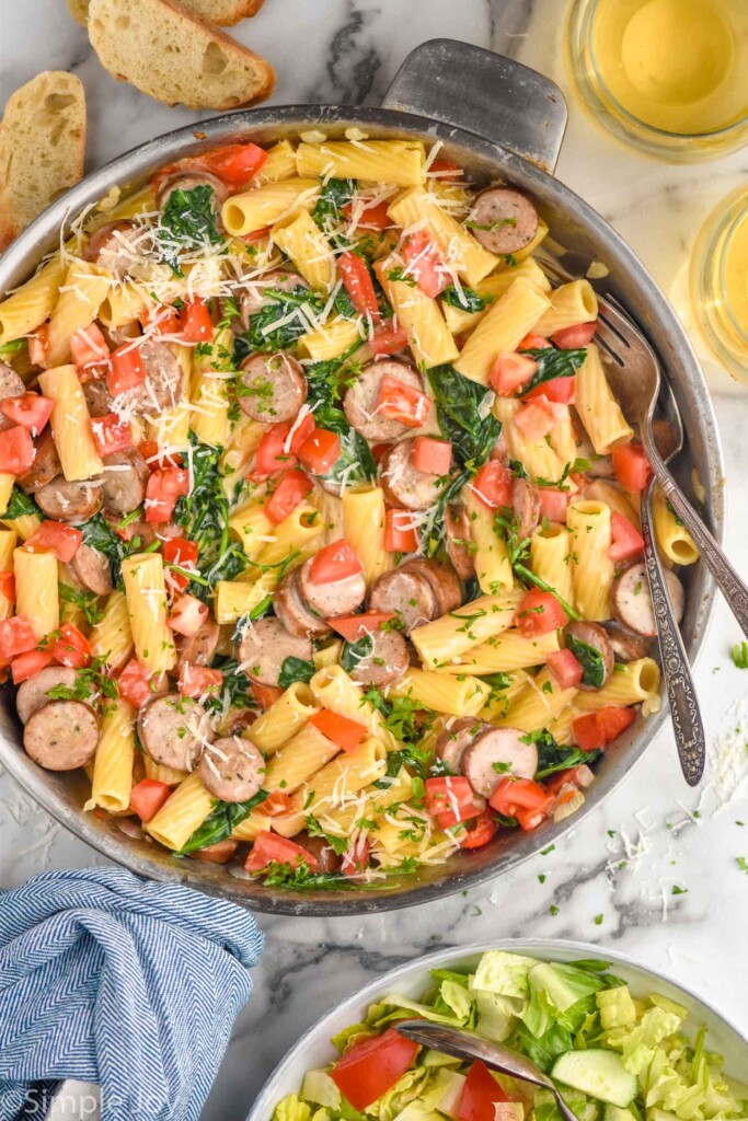 overhead of large skillet of One Pot Sausage Pasta topped with freshly grated parmesan cheese and parsley, bowl of salad, two glasses of white wine, and slices of bread surrounded