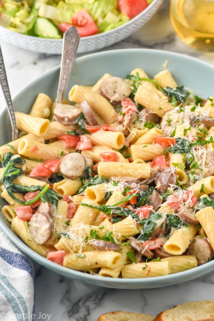 bowl of One Pot Sausage Pasta topped with freshly grated parmesan cheese and parsley. Bowl of salad and glass of white wine sitting in background