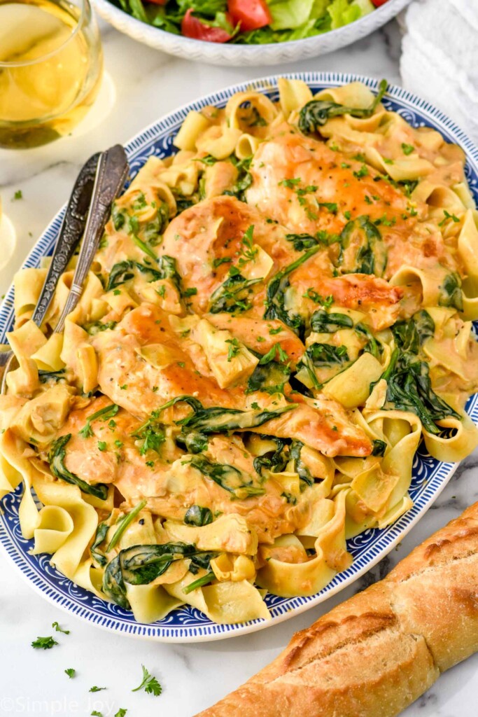 platter of Spinach Artichoke Chicken with pasta and two forks for serving. Bowl of salad and baguette sit beside