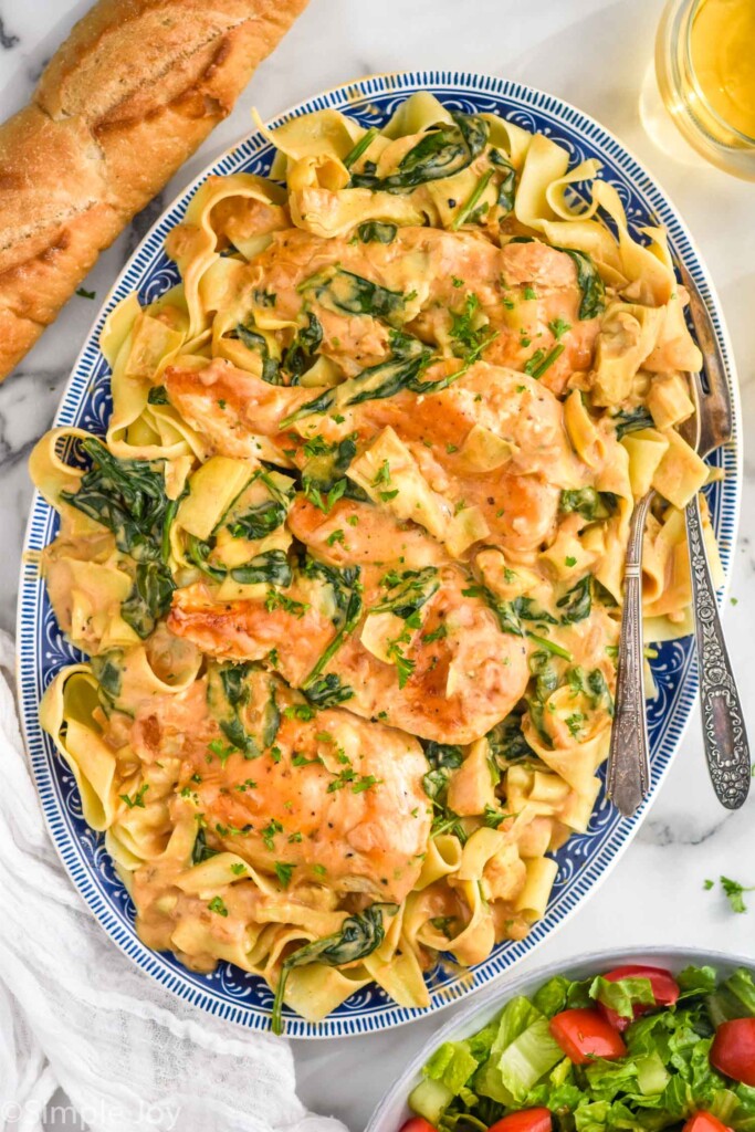 overhead of platter of Spinach Artichoke Chicken with pasta and two forks for serving. Bowl of salad and baguette sit beside