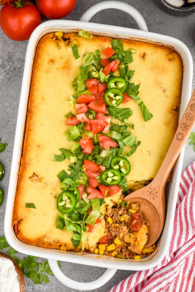 overhead of casserole dish of Tamale Pie topped with lettuce, diced tomatoes, and jalapenos. Wooden spoon in dish for serving.