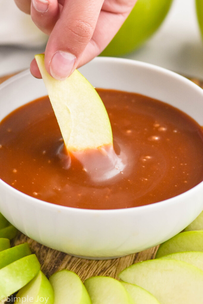 hand holding apple slice dipped in bowl of caramel apple dip. Apple slices surrounding bowl