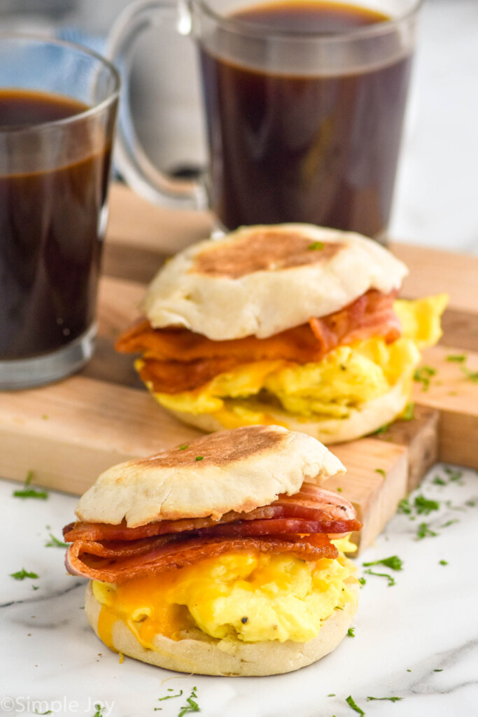 Two Breakfast Sandwiches with bacon, eggs, and cheese on a toasted english muffin with two mugs of coffee sitting in background