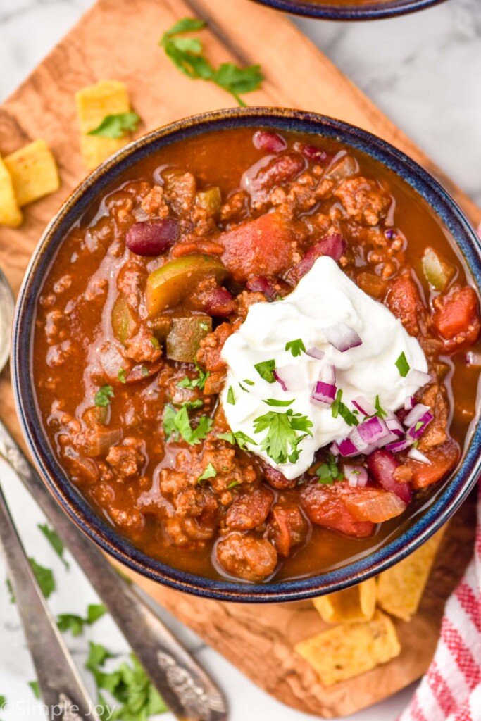 overhead of bowl of chili topped with sour cream, diced red onion, and fresh parsley with corn chips sitting beside bowl