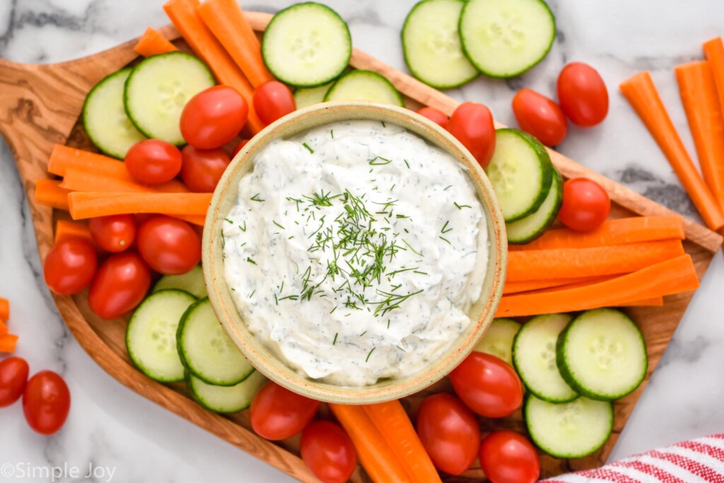 overhead of board of sliced cucumbers, carrot sticks, cherry tomatoes, and bowl of ranch dip topped with dill