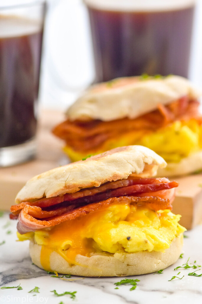 A Breakfast Sandwich with bacon, eggs, and cheese on an english muffin. Breakfast Sandwich and two mugs of coffee sitting in background