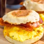 Breakfast Sandwich with slices of bacon, eggs, and cheese on an english muffin. Breakfast Sandwich and mugs of coffee sitting in background