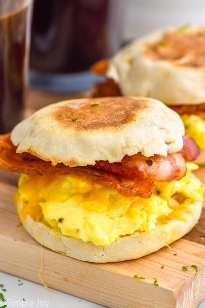 Breakfast Sandwich with slices of bacon, eggs, and cheese on an english muffin. Breakfast Sandwich and mugs of coffee sitting in background