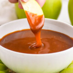 hand holding apple slice dipped in bowl of caramel apple dip. Apple slices surrounding bowl