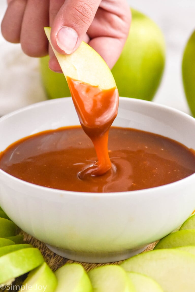 hand holding apple slice dipped in bowl of caramel apple dip. Apple slices surrounding bowl