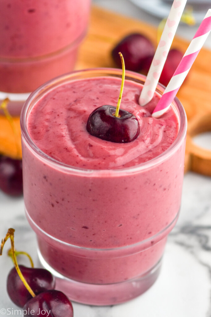 angled view of a cherry smoothie in a glass with a cherry on top and two paper straws