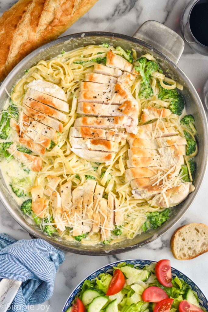 Overhead of skillet of pasta with alfredo sauce and broccoli topped with sliced chicken. Bowl of salad and baguette sitting beside.