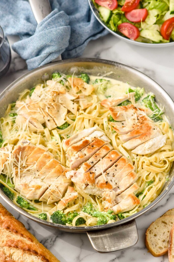 Skillet of chicken and broccoli alfredo with shredded parmesan cheese on top. Bowl of salad and sliced baguette sitting beside