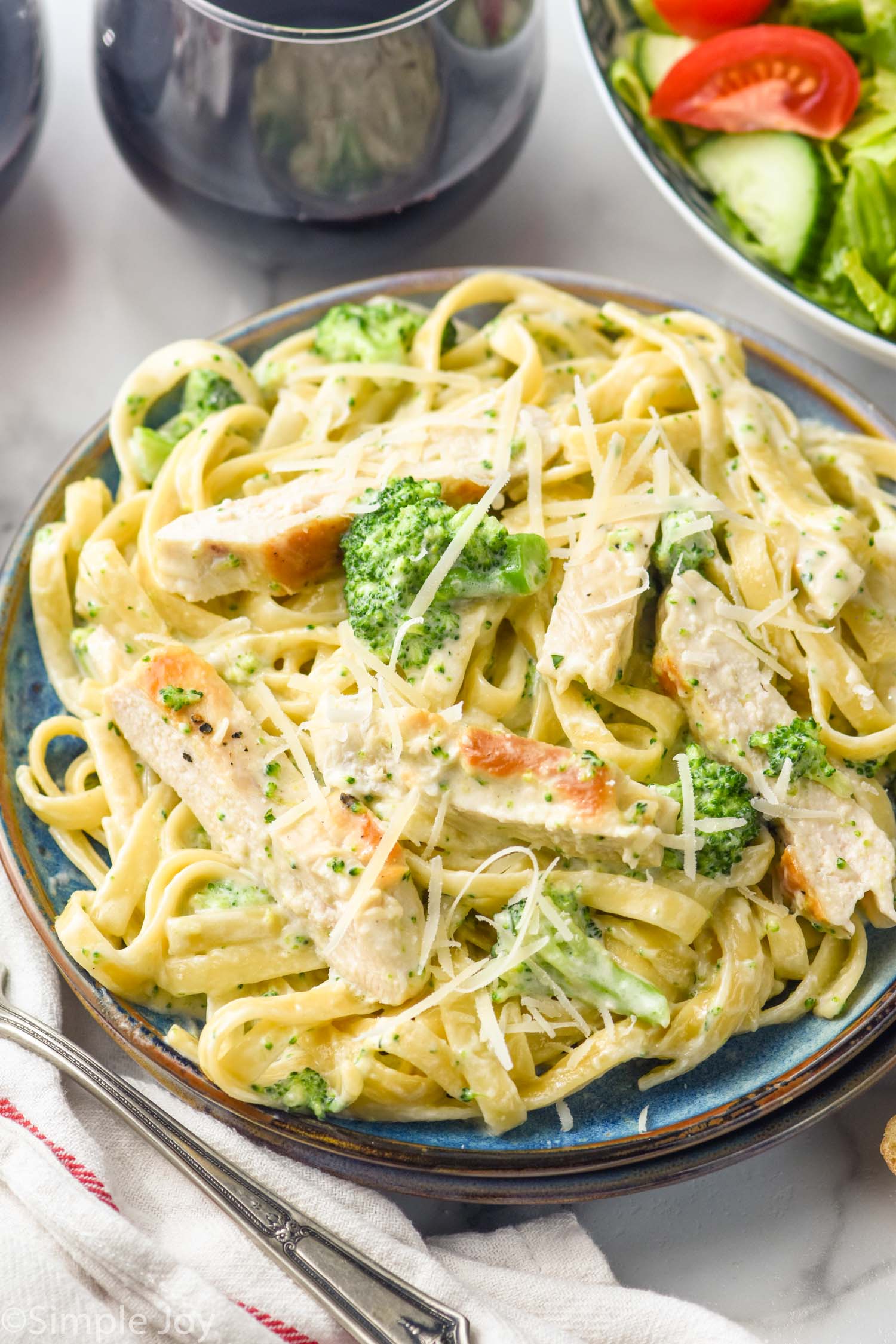 plate of Chicken Broccoli Alfredo topped with shredded parmesan cheese. Glass of wine and bowl of salad sitting beside