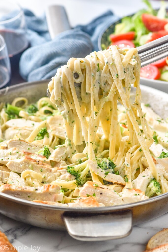 tongs pulling fettuccini pasta will alfredo sauce out of skillet of Chicken Broccoli Alfredo