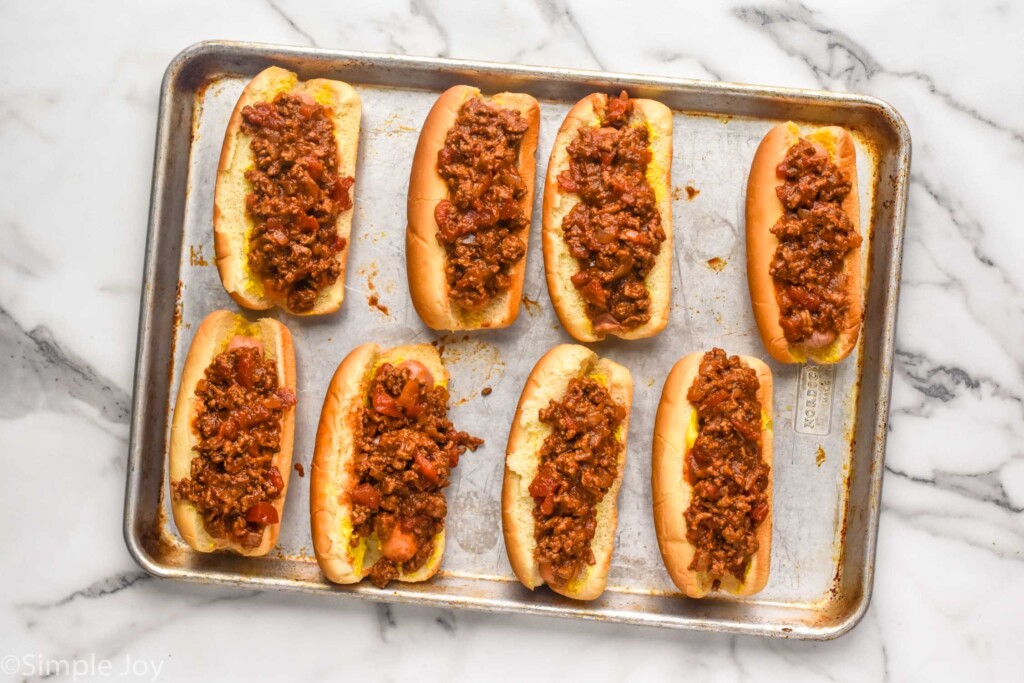 overhead of baking sheet with hot dog buns, hot dogs, topped with chili to make Chili Dogs