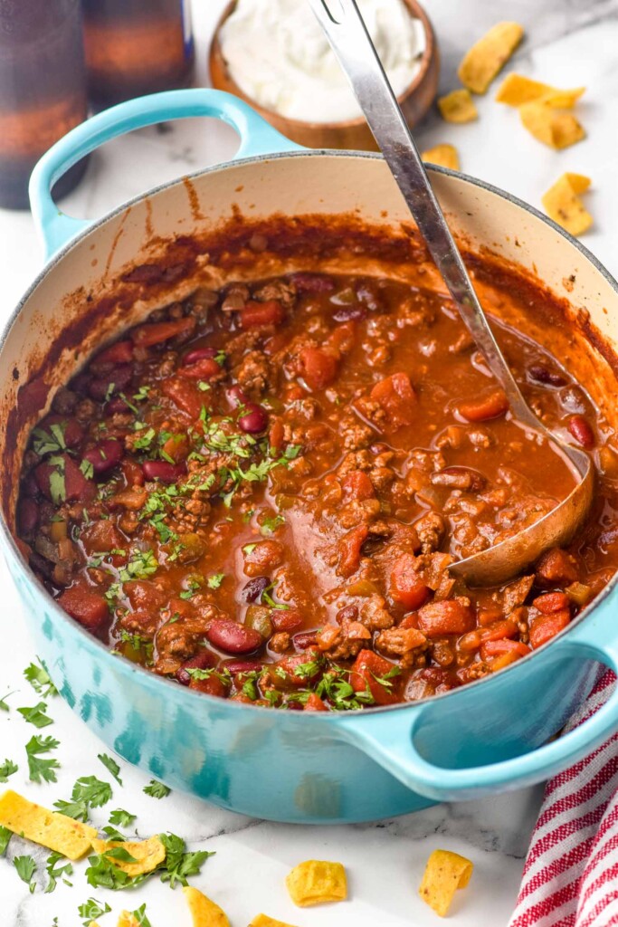 large pot of chili with fresh parsley and a ladle with corn chips and parsley sitting beside.