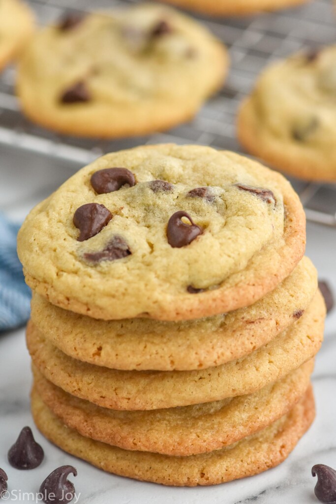 Stack of Chocolate Chip Cookies with chocolate chips in front and Chocolate Chip Cookies on a wire cooling rack sitting behind