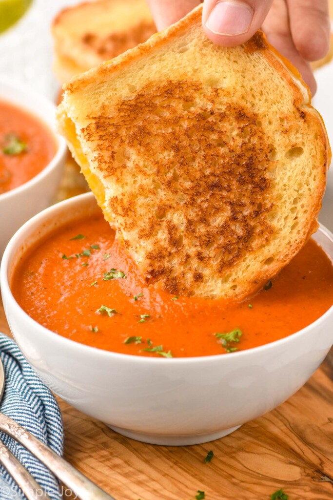 Person's hand dipping grilled cheese sandwich into a bowl of Tomato Soup garnished with parsley.