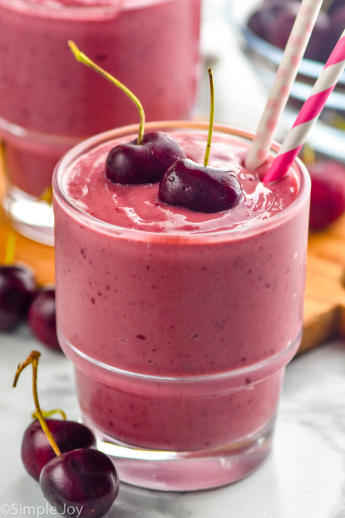 a glass of an easy cherry smoothie with two stemmed cherries on top and paper straws sticking out