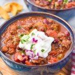 two bowls of chili topped with sour cream, diced red onion, and fresh parsley. Corn chips siting beside