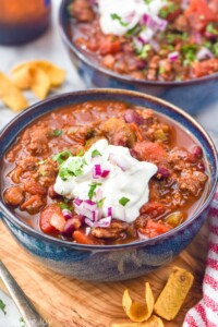 two bowls of chili topped with sour cream, diced red onion, and fresh parsley. Corn chips siting beside