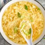 hand holding spoon of Egg Drop Soup over a bowl of Egg Drop Soup topped with diced green onions