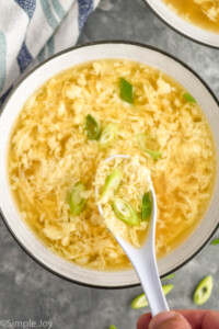 hand holding spoon of Egg Drop Soup over a bowl of Egg Drop Soup topped with diced green onions