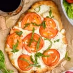 overhead of Flatbread Pizza topped with tomato slices and fresh basil. Two glasses of red wine sitting beside